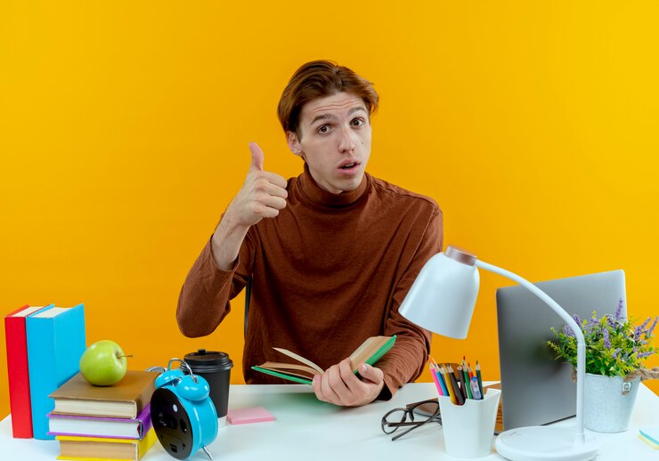 thinking young student boy sitting desk with school tools holding book his thumb up yellow 141793 74786