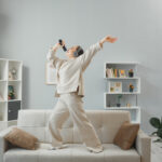 Liburan di Rumah Bisa Ngapain Aja Ya? 6 happy young woman casual clothing with headphones dancing couch home interior holding remote using it as microphone singing having fun