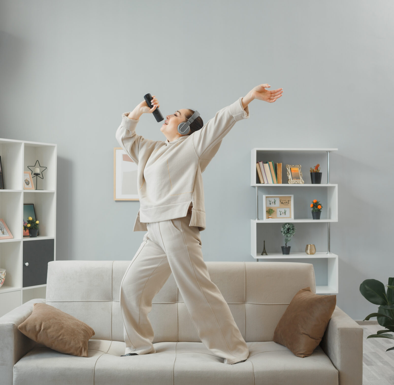 happy young woman casual clothing with headphones dancing couch home interior holding remote using it as microphone singing having fun