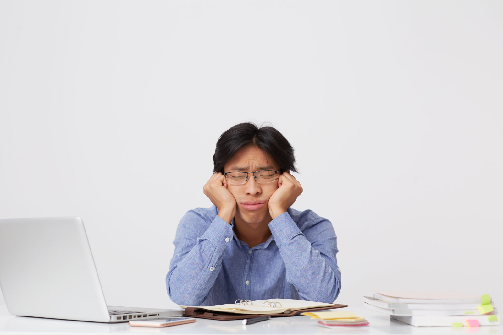 tired sleepy asian young business man glasses with head hands sitting sleeping workplace table white wall