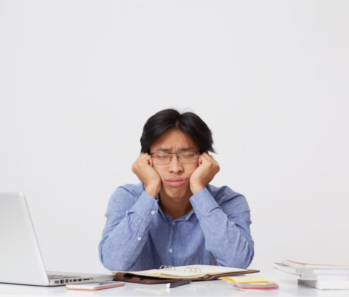 tired sleepy asian young business man glasses with head hands sitting sleeping workplace table white wall
