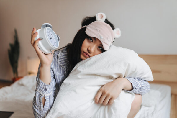 sad girl pajamas sleep mask is hugging white pillow holding alarm clock