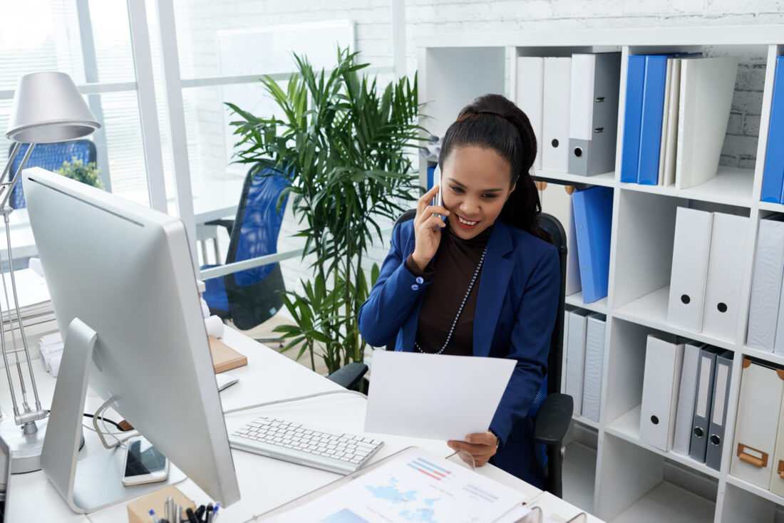 smiling asian woman sitting desk office looking document talking mobile phone