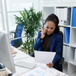 smiling asian woman sitting desk office looking document talking mobile phone