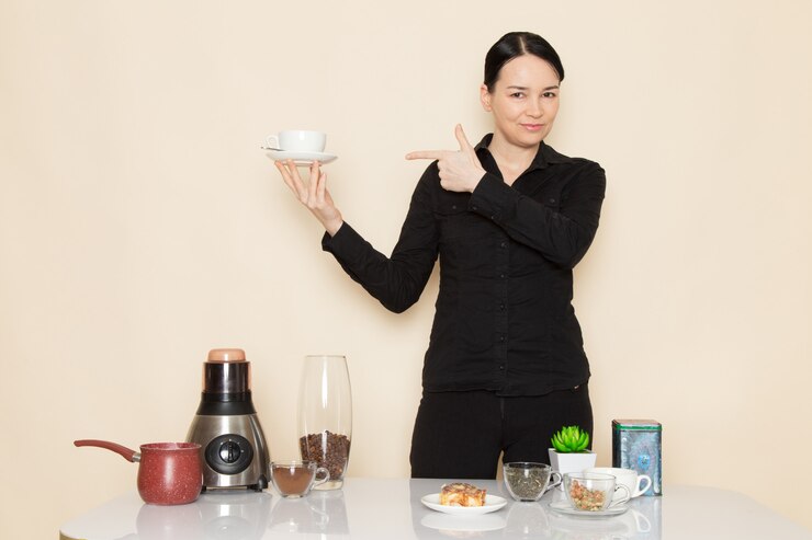 female barista black shirt trousers front table with coffee ingredients equipment white wall 140725 11636